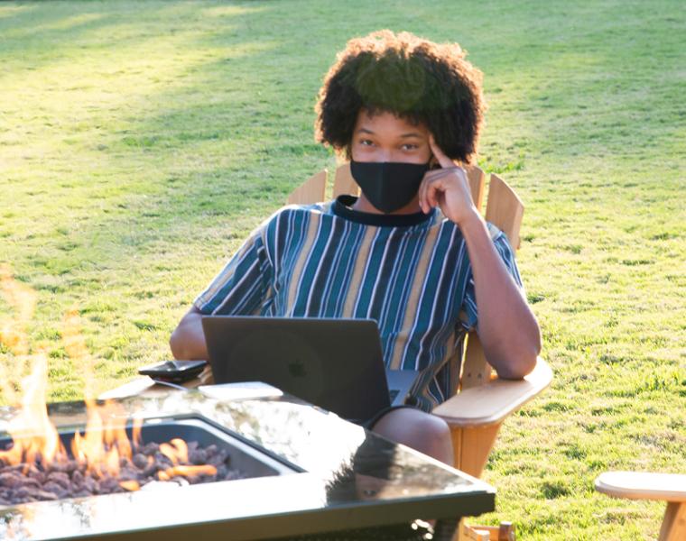 student wearing a mask studying with a laptop sitting by a fire pit