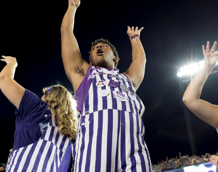 students dressed in purple and white striped overalls, cheering at football game
